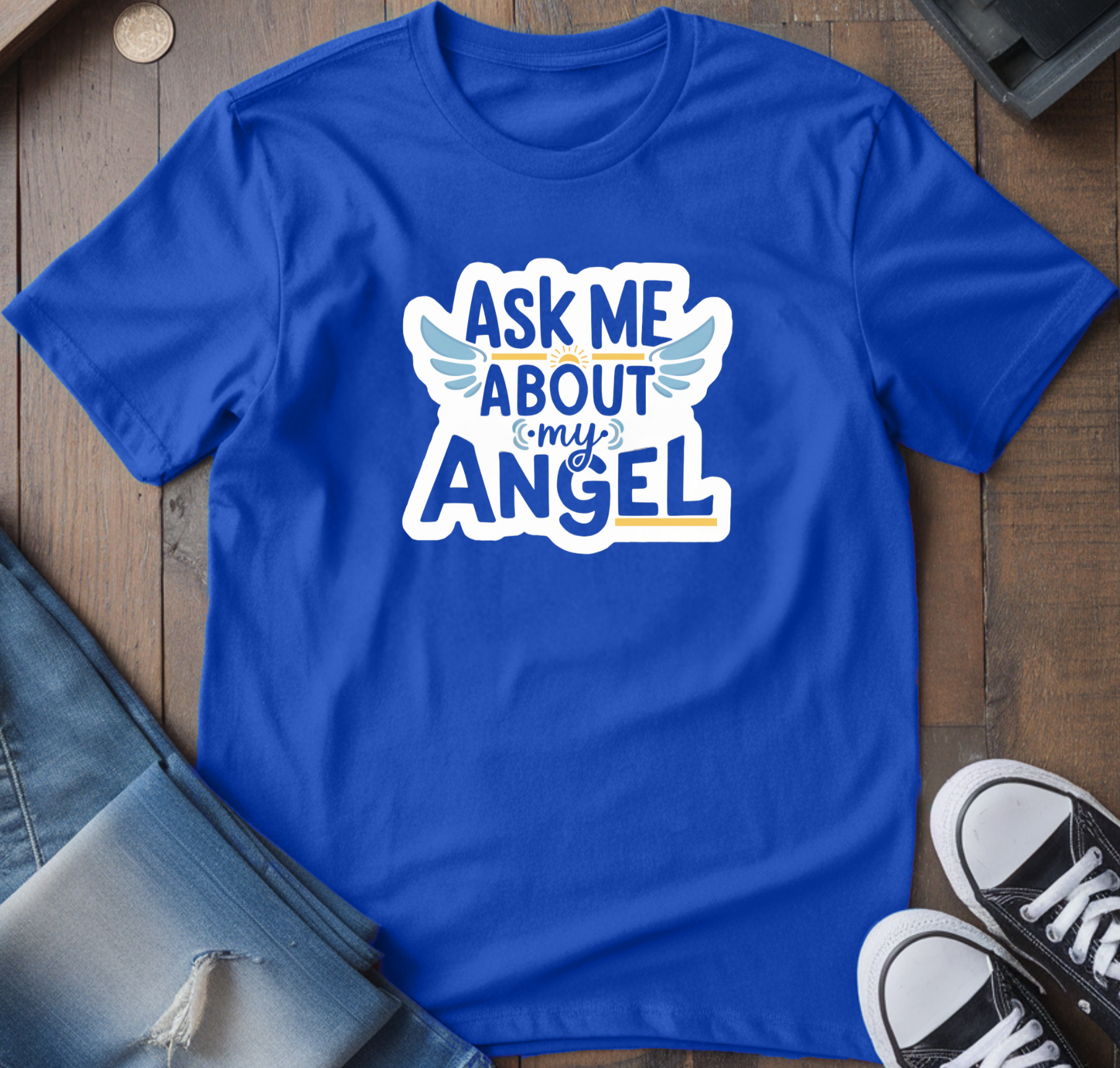 Blue t-shirt with 'Ask Me About My Angel' design on a wooden surface with jeans and sneakers.