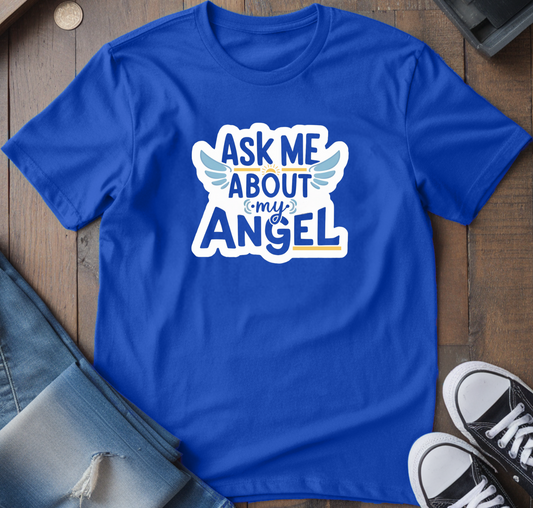 Blue t-shirt with 'Ask Me About My Angel' design on a wooden surface with jeans and sneakers.