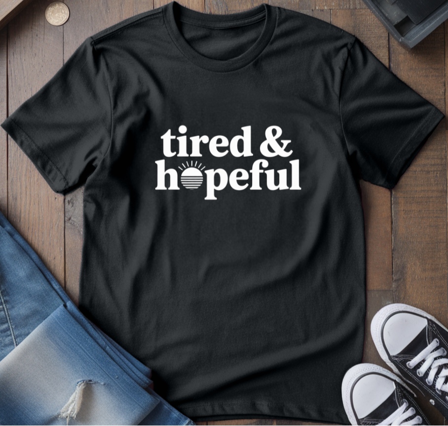 Black t-shirt with 'tired & hopeful' text, blue jeans, and black sneakers on a wooden surface