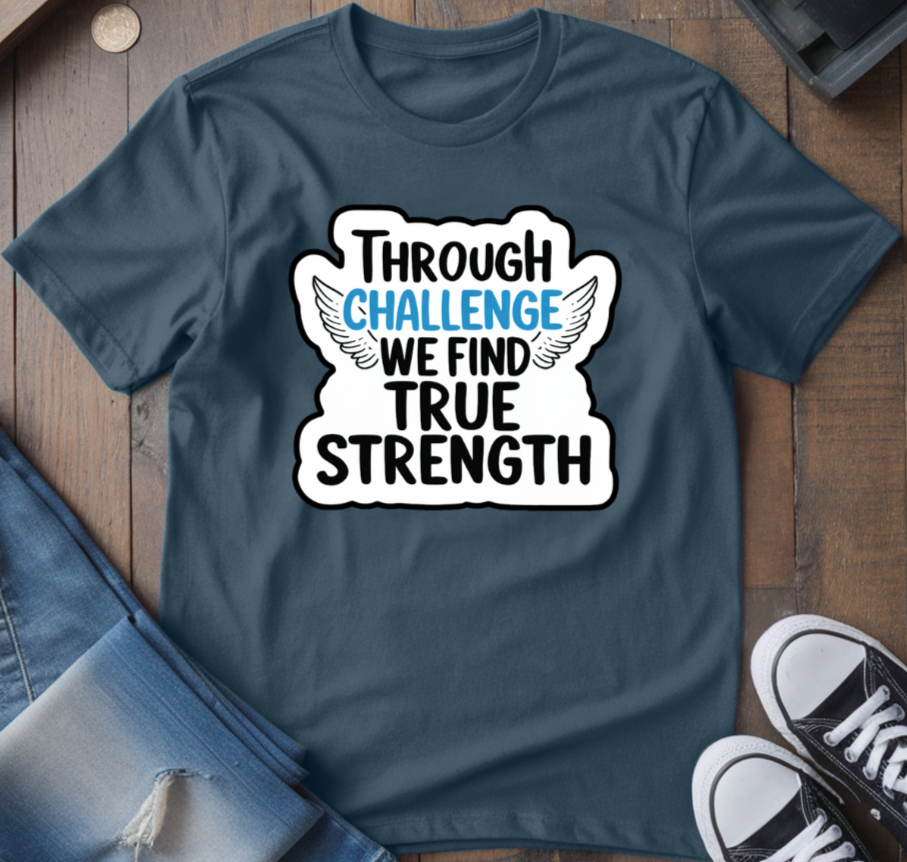 A navy blue crew neck t-shirt with the text 'THROUGH CHALLENGE WE FIND TRUE STRENGTH' printed on the front, displayed flat with jeans and sneakers on a wooden surface.