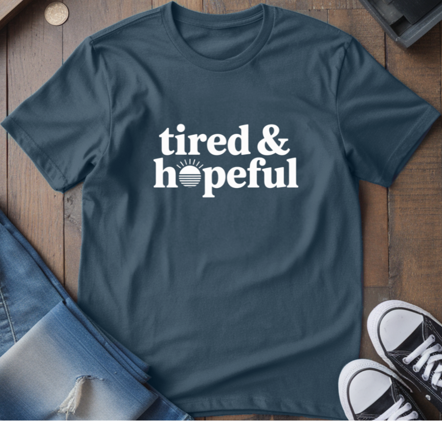 Dark Heather t-shirt with 'tired & hopeful' text, paired with jeans and sneakers on a wooden surface.