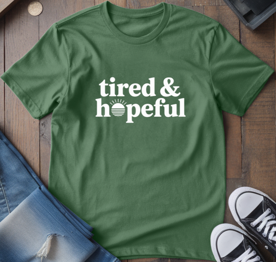 Green t-shirt with 'tired & hopeful' text, paired with jeans and sneakers on a wooden surface.