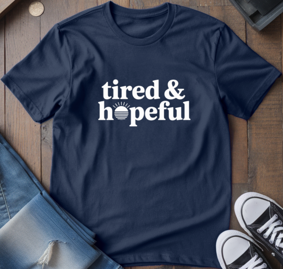 Navy blue t-shirt with 'tired & hopeful' text on a wooden surface with jeans and sneakers.