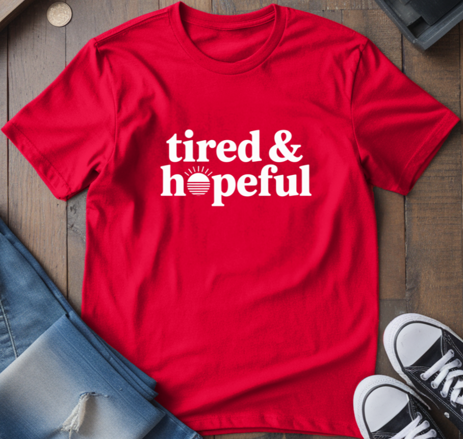 Red t-shirt with 'tired & hopeful' text on a wooden surface with jeans and sneakers.