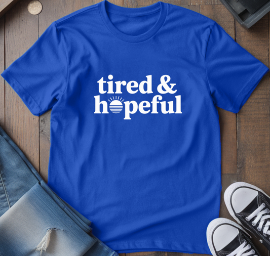 Blue t-shirt with 'tired & hopeful' text on a wooden surface with jeans and sneakers.