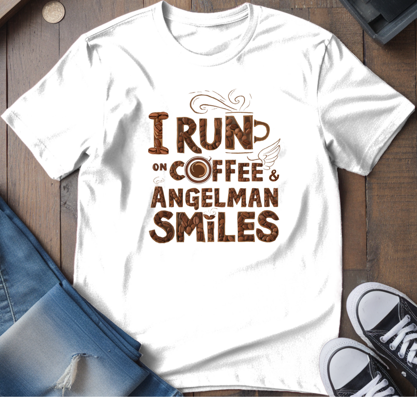 White crew neck t-shirt with the text 'I RUN ON COFFEE & ANGELMAN SMILES' printed on the front, presented on a wooden surface accompanied by jeans and sneakers.